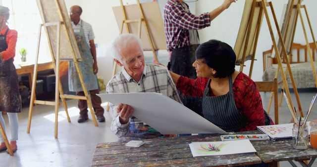 Senior man enjoying interactive art class with attentive female teacher in studio setting. Great for themes involving lifelong learning, creativity in later years, artistic mentorship, educational workshops, and intergenerational activities.