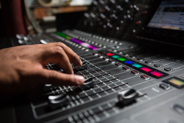 Hand of male audio engineer adjusting sound mixer in recording studio. Ideal for use in articles about music production, sound engineering, professional audio equipment, and the music industry. Suitable for illustrating concepts related to studio work, audio technology, and sound control.