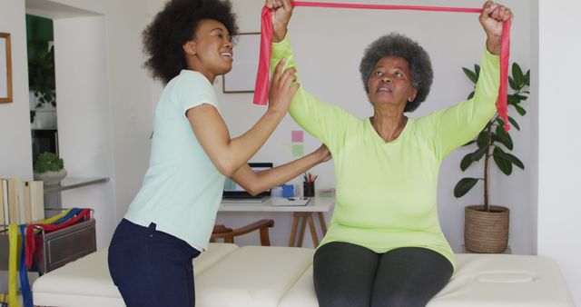 Senior woman sitting on therapy table, using resistance band for exercise. Young therapist assisting and guiding her movements. Ideal for fitness programs for seniors, physical therapy guides, elderly care resources, and promoting active lifestyles among older adults.