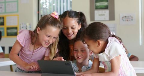 Elementary girls leaning around tablet, learning and collaborating, smiling in classroom