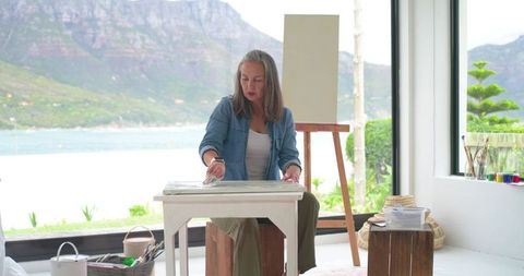 Mature woman painting by coastal window in sunlit cozy studio with easel and art supplies