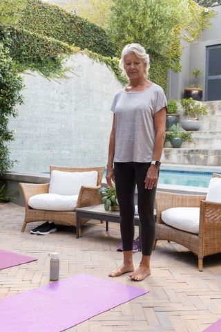 Senior woman practicing barefoot yoga on patio by pool with purple mats and smartwatch