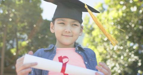 Smiling schoolgirl wearing mortarboard and holding diploma with yellow tassel outdoors