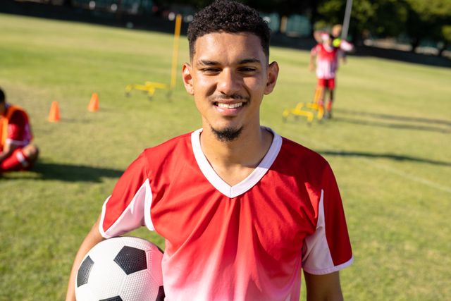 Portrait of caucasian confident player wearing red jersey and holding ball standing in playground. Summer, unaltered, sport, soccer and competition concept.