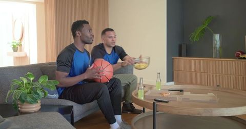 Black and latino friends watching basketball on couch holding basketball sharing snacks