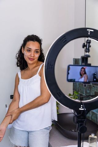 Woman applying skincare while filming with ring light and smartphone at bathroom vanity