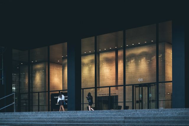 This scene captures a modern office building with a sleek glass facade during the evening. Two people can be seen walking near the entrance, adding a professional and dynamic feel. This image is ideal for editorial use in business-related contexts, corporate websites, architectural portfolios, or urban lifestyle magazines.