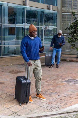 African american travelers using smartphones waiting outside glass building with luggage