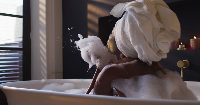 African american woman with towel taking bath and blowing foam in bathroom. health and beauty concept.