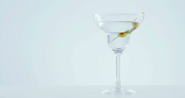 A martini glass with a clear liquid and an olive garnish stands against a light background, with copy space. Its elegant presentation suggests a sophisticated setting or a concept related to leisure and luxury.