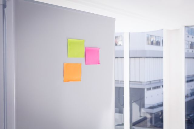 Colorful sticky notes are attached to a refrigerator in a modern kitchen. The notes are bright green, pink, and orange, adding a pop of color to the sleek, minimalist space. This image can be used for themes related to home organization, planning, reminders, and modern kitchen interiors.