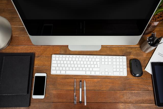 Neatly arranged office desk featuring a computer, mobile phone, keyboard, mouse, and pens on a wooden surface. Ideal for illustrating modern workspaces, business environments, and productivity themes. Suitable for use in articles, blogs, and presentations about office organization, technology in the workplace, and professional settings.