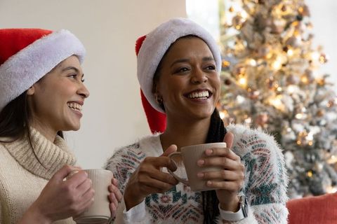 Multiracial friends in santa hats holding mugs, enjoying cozy christmas living room