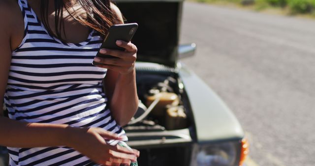 African american woman using smartphone while standing near her broken down car on road. road trip travel and adventure concept