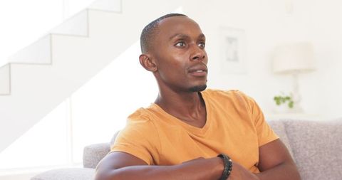 Contemplative man sitting on sofa gazing upward wearing orange tee and beaded bracelet