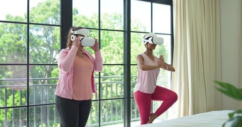 Two women practicing vr yoga at home, balancing in tree pose with virtual reality headsets