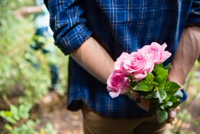 Man holding pink roses behind his back, creating a sense of anticipation and surprise. Ideal for use in romantic, relationship, and nature-themed projects. Perfect for illustrating love, surprise gestures, and outdoor activities.