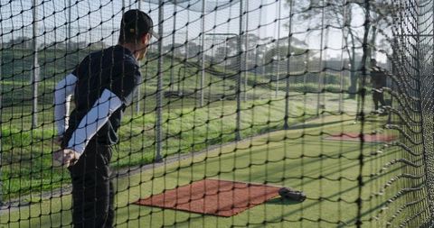 Two baseball players practicing in outdoor batting cage with pitching mats, glove, netting