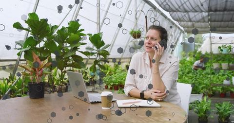 Mature woman managing greenhouse office talking on smartphone with laptop and plants