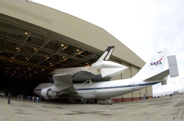 <i>[Photo courtesy of Boeing photographer Bob Williams.]</i> The orbiter Columbia, atop a modified Boeing 747, rolls under protective cover at Palmdale, Calif. Columbia has been undergoing modifications and upgrades at Boeing’s Orbiter Assembly Facility in Palmdale and is ready to return to Kennedy Space Center. Ferry preparations and the flight plan are contingent upon weather conditions in California and enroute to Florida.