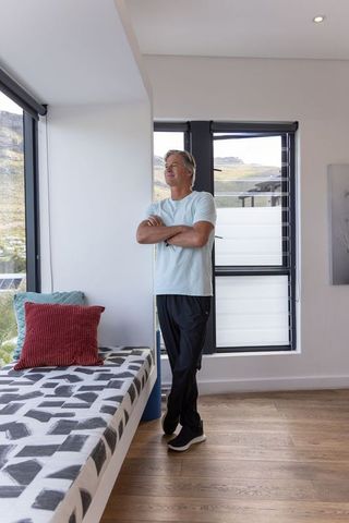 Mature man standing by window in sunlit modern minimalist interior with geometric daybed