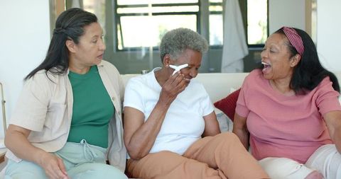 Multigenerational diverse women laughing and comforting each other on sunlit sofa