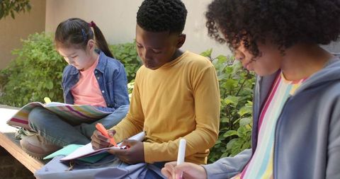 Diverse children studying outdoors on bench using notebooks and markers for schoolwork