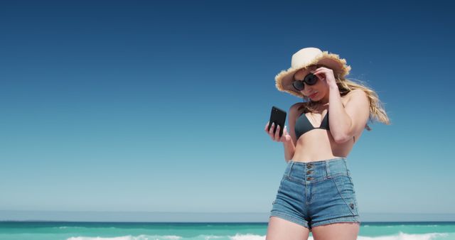 Young woman stands on beach wearing denim shorts and straw hat, holding phone, looking at screen. Ocean waves and clear blue sky in background. Ideal for promoting summer fashion, beach vacations, leisure activities, and smartphone use outdoors.