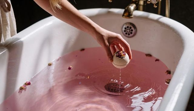 Hand holding dropping rose petal bath bomb into pink water vintage tub luxury brass faucet
