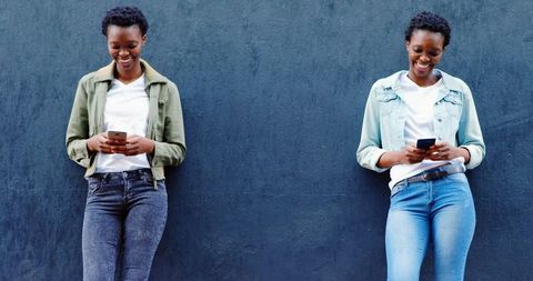 Twin sisters using smartphones leaning against urban wall