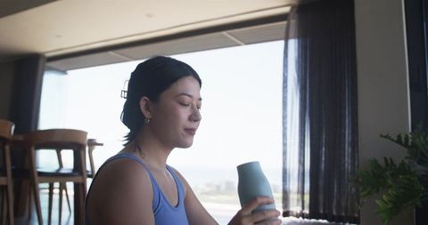 Woman sitting holding teal tumbler by coastal window in modern minimalist condo serene calm