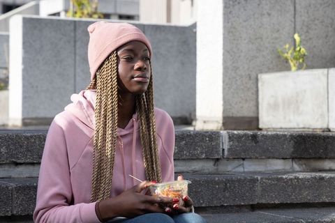 African american woman sitting on city steps holding salad container wearing pink beanie