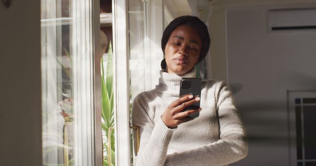 Pensive African Woman Standing by Window Using Smartphone in Natural Light - Download Free Stock Images Pikwizard.com