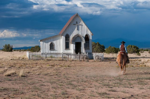 Cowboy riding horse along dirt path towards an isolated, abandoned church on a vast desert landscape. Blue sky with dramatic clouds rolls in background. Perfect for themes involving western lifestyle, rural solitude, historical adventure, travel, and American Southwest culture.