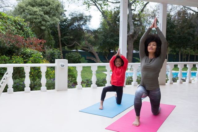 Image shows mother and daughter practicing yoga on their porch, surrounded by greenery. Suitable for use in articles or content about family fitness, healthy lifestyle, home workouts, or parent-child bonding activities.