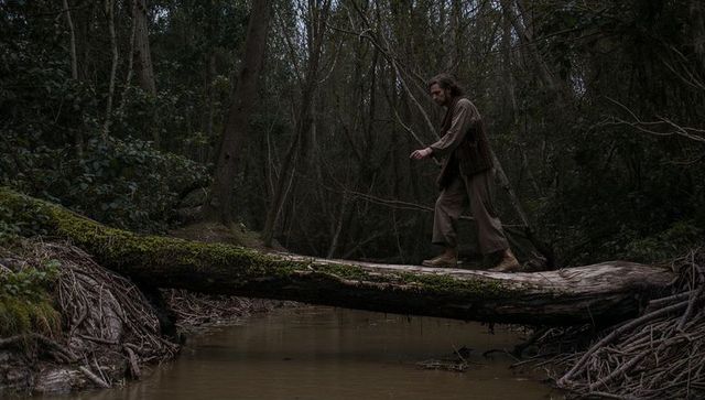 Solitary man balancing on mossy log over muddy creek walking through moody woodland autumn