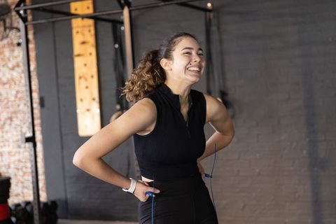 Energetic smiling woman holding blue jump rope in gym wearing black zip top and leggings