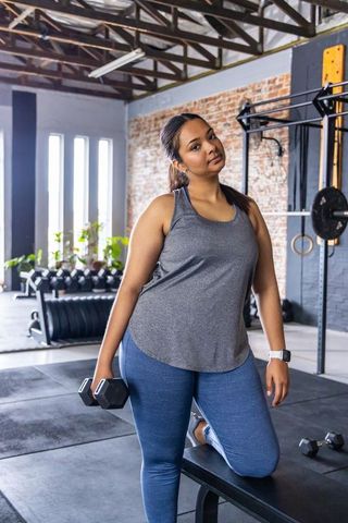 Asian woman training with dumbbell kneeling on bench in industrial gym daylight
