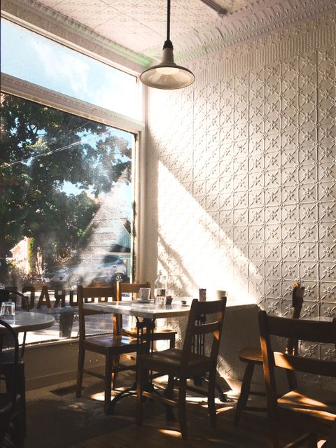 Shows warm morning light flooding through window into a charming coffee shop with empty wooden chairs and tables. Ideal for illustrating cozy cafe atmospheres, vintage interior design, morning routines, relaxed public spaces, coffee commendations.