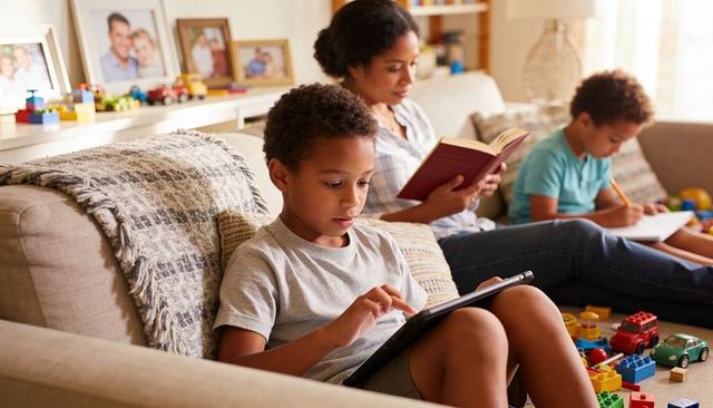 Family relaxing at home children using tablet and drawing while parent reading on sofa