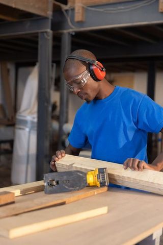 African American Carpenter Inspecting Wooden Plank Beside Power Planer in Busy Woodshop