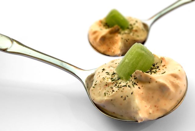 Up-close view of a gourmet appetizer consisting of a creamy pink dip elegantly served on metal spoons, each topped with a slice of green celery and sprinkled with herbs. Ideal for use in food blogs, restaurant menus, or culinary magazines to illustrate elegant food presentation, appetizer ideas, or detailed food arrangements.