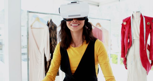 Woman wearing VR headset and smiling while exploring virtual reality shopping experience in a fashion store. Clothing and outfits visible in the background. Ideal for topics related to technology in retail, futuristic shopping experiences, and joy of modern innovations. Useful for illustrating concepts in tech advancements in fashion retail and customer engagement strategies.