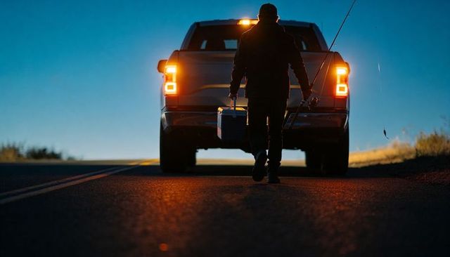 Twilight angler walking toward pickup carrying cooler and rod backlit by taillights