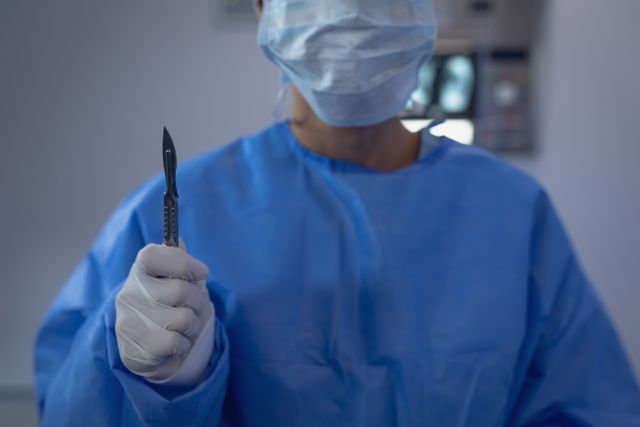 Male surgeon in blue scrubs holding a scalpel in a hospital operating room. Suitable for use in medical, healthcare, and surgical procedure contexts, as well as educational materials and healthcare advertisements.