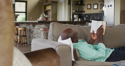 Reclining senior man reading paperback on sofa in open-plan home living room cozy daylight