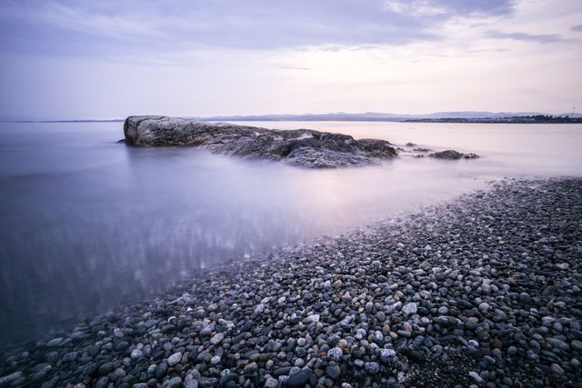 This photo captures a tranquil moment on a pebble beach at dusk, with calm waters and an overcast sky creating a serene mood. Perfect for promoting relaxation, travel, coastal living, or environmental awareness. Ideal for websites, brochures, or social media posts looking to evoke a sense of peace and natural beauty.