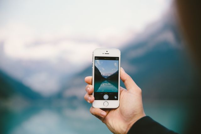 Hand holding smartphone while taking photo of scenic mountain lake backdrop. Useful for travel advertisements, nature blogs, technology features, and content about outdoor activities and photography tutorials.