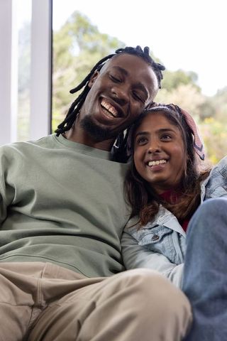 Diverse couple sitting on couch by window, leaning and smiling, cozy intimate home moment