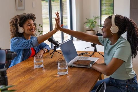 African american podcast co-hosts recording, high-fiving and smiling in sunlit home studio